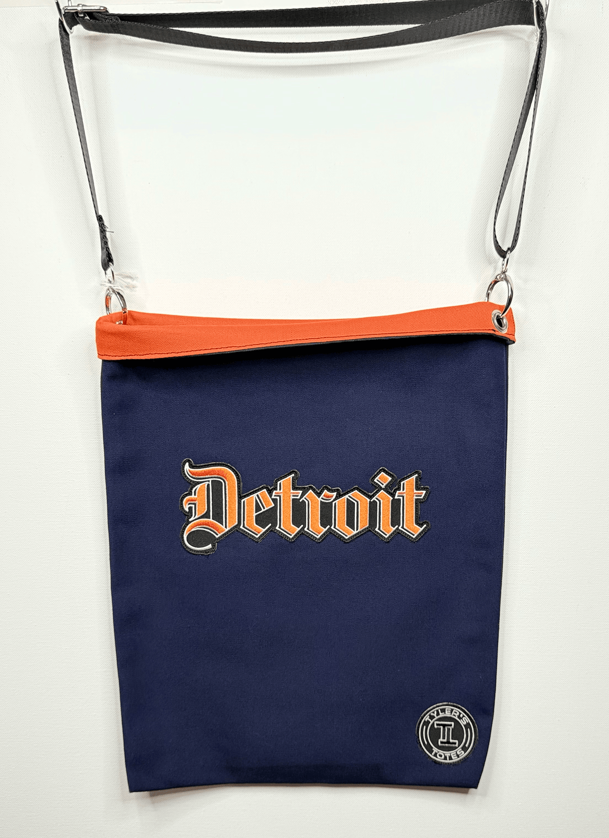 A navy blue Detroit crossbody bag with foldable brim, featuring the word 'Detroit' in orange. It has a black, adjustable shoulder strap and is displayed against a plain white background.